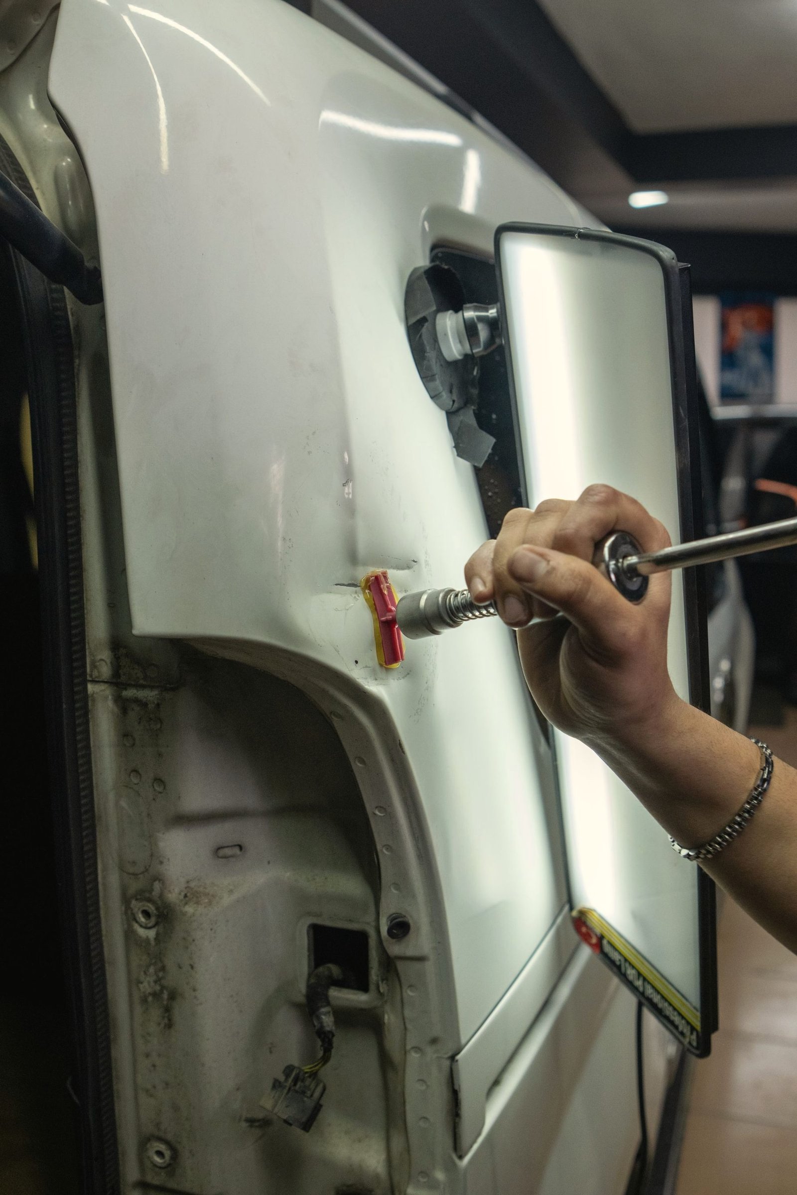 A hand tool pulling a dent out of a white door panel under a striped light