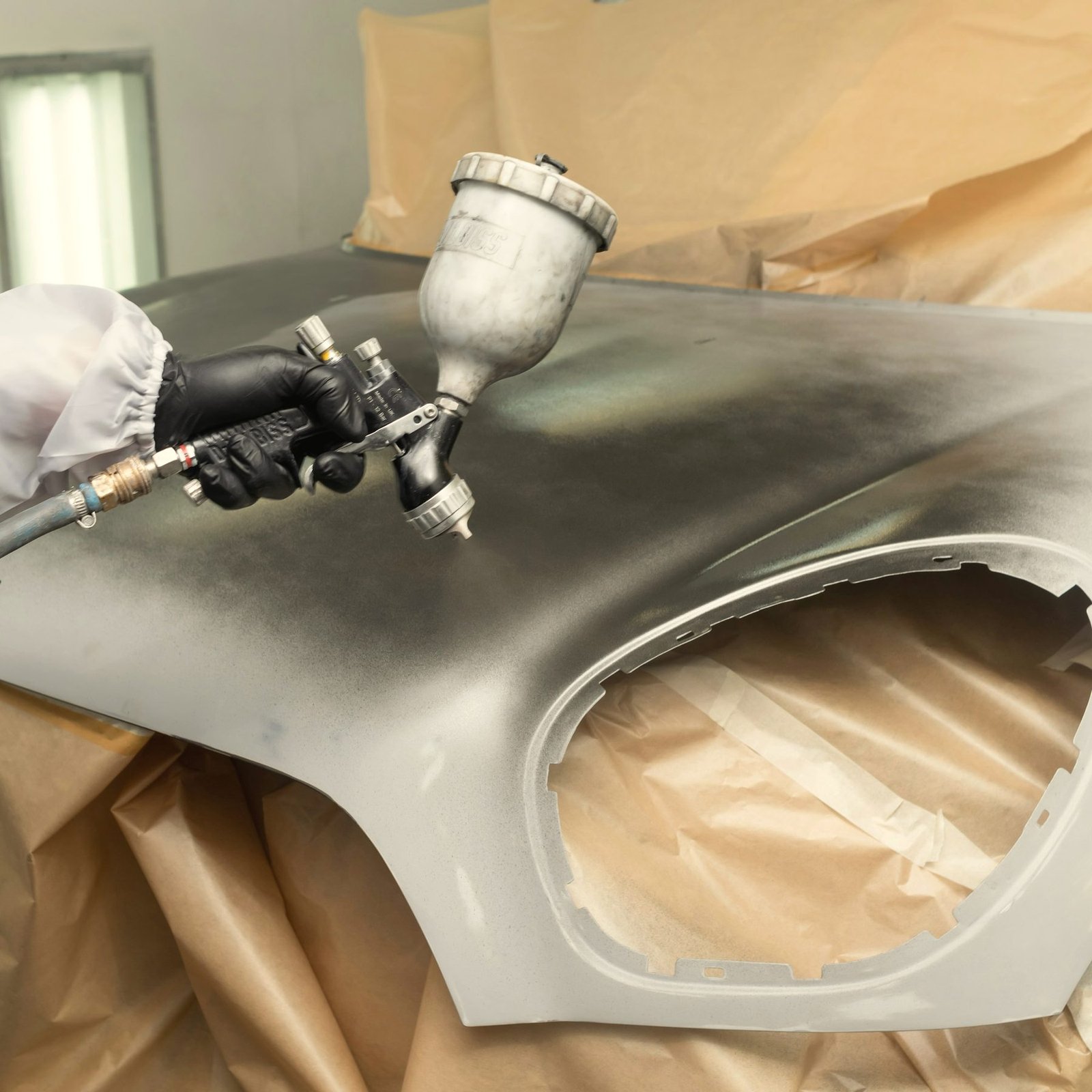 A spray gun laying silver paint on a fender, with tan masking paper taped around it
