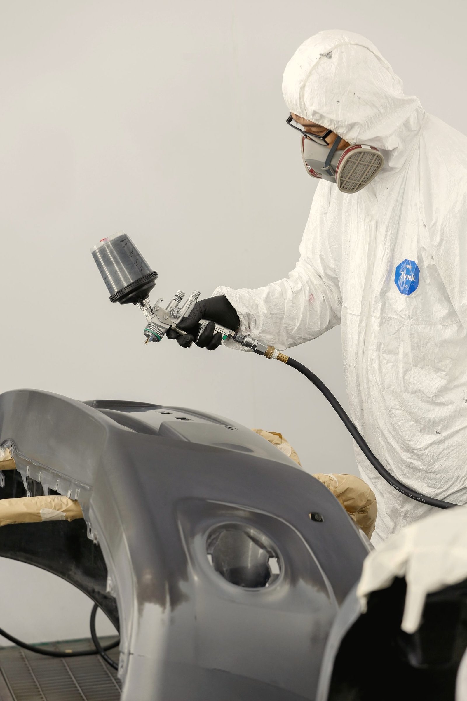 A painter in a white suit and respirator spraying a bumper cover